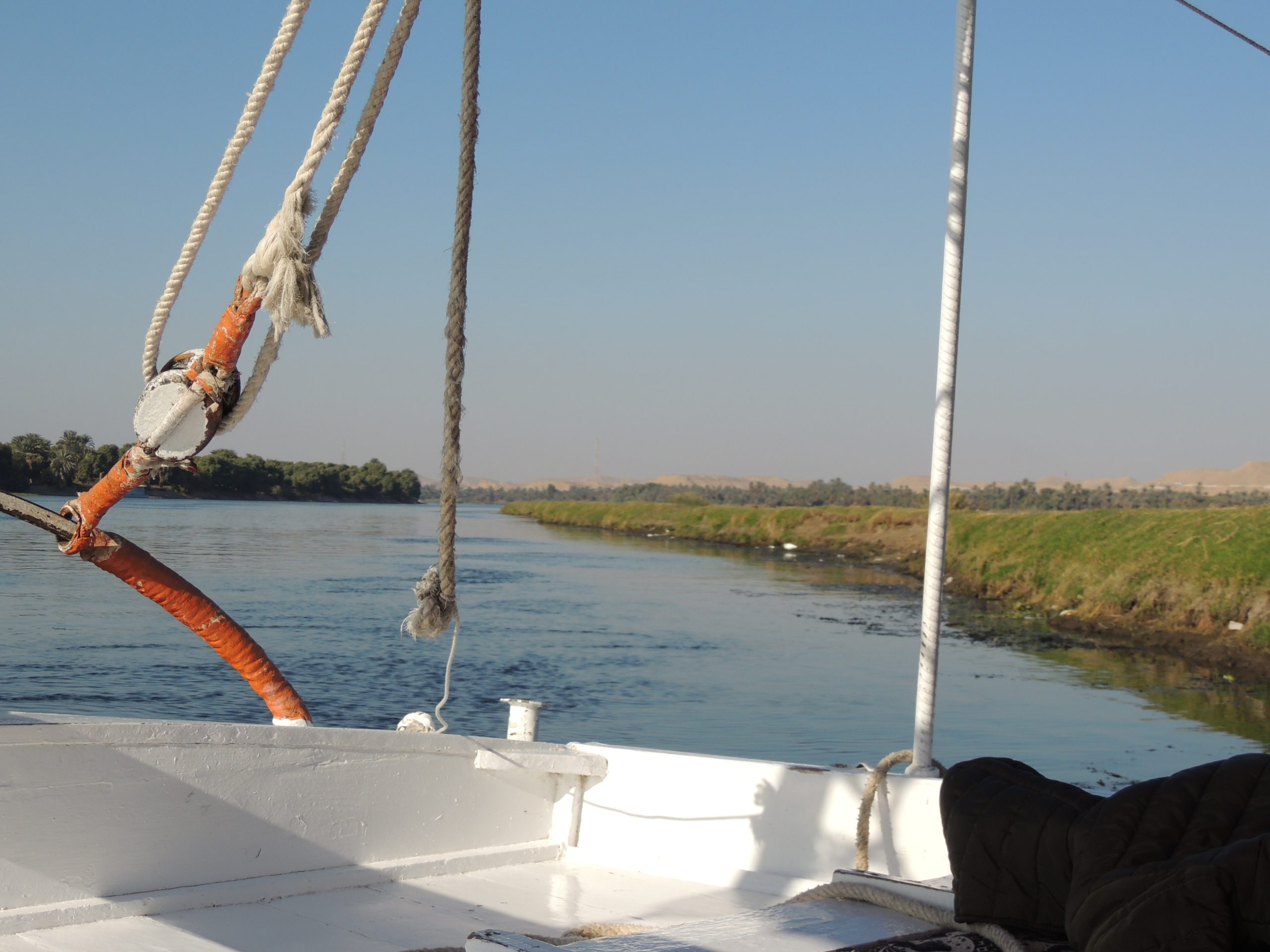 View from a traditional Egyptian felucca sailing along the Nile River with green riverbanks and desert landscape in Upper Egypt