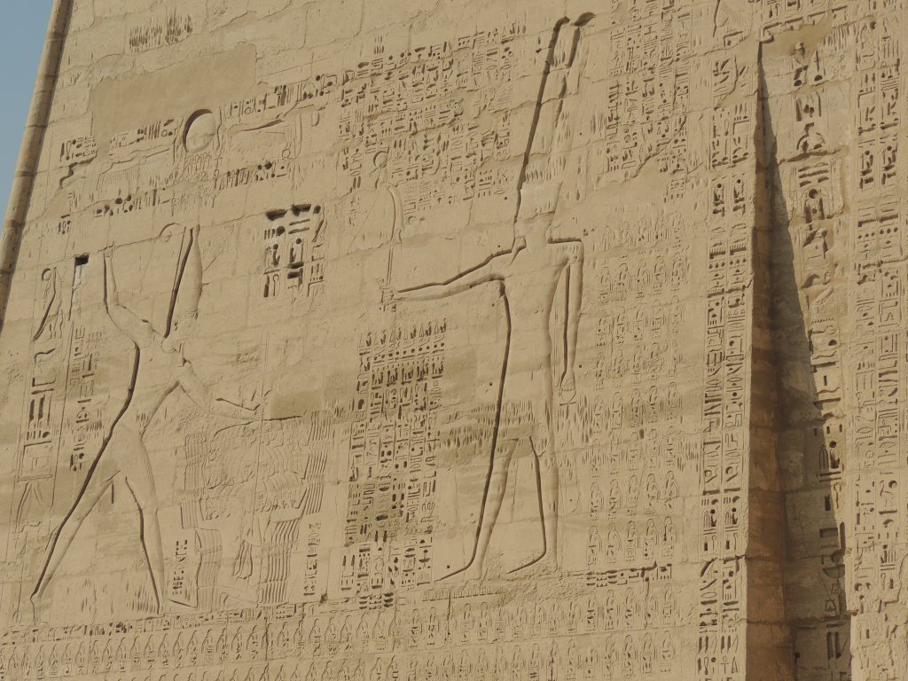 Stone wall relief showing an ancient Egyptian pharaoh wearing a tall crown performing a ritual offering scene carved in a temple wall in Upper Egypt.